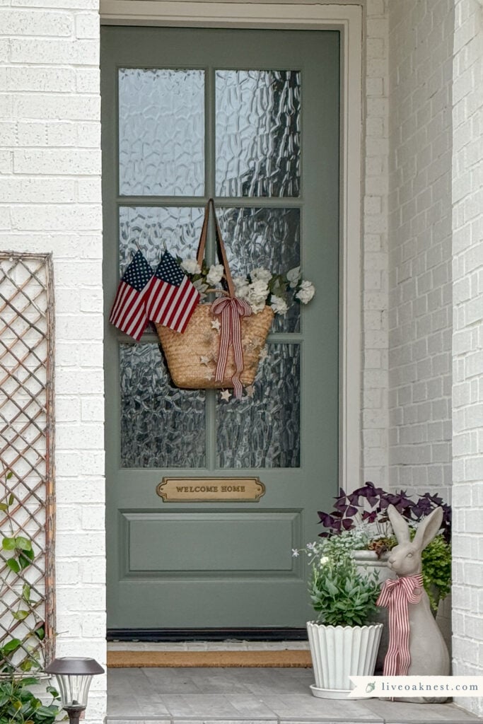Sage Green Front Door, Basket of Flowers with American Flags, Star Garland, Red and White Ribbon. Porch Plants with Standing Rabbit Figurine with Red and White Patriotic Ribbon around Neck